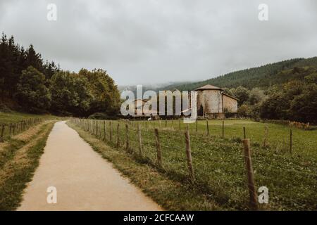 Narrow path near fenced lawn surrounded by forest on hillside and old buildings under cloudy sky in afternoon in countryside Stock Photo