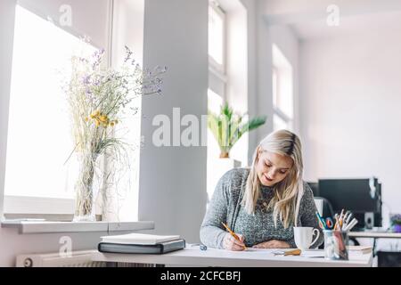 Smiling blond Woman doing linearity and first sketches at table in light office near window Stock Photo