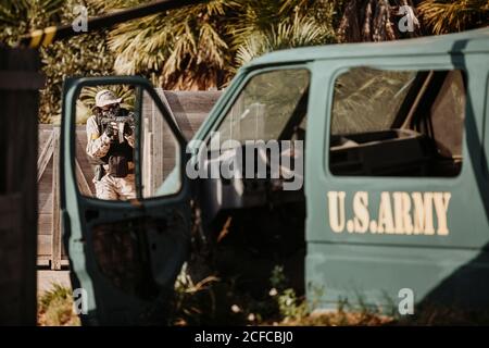 man in camouflage hiding behind wooden barrier and shooting airsoft gun behind military vehicle while playing tactical game Stock Photo