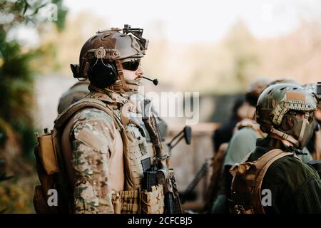 Side view of adult man in military uniform and with airsoft gun listening to briefing with squad Stock Photo