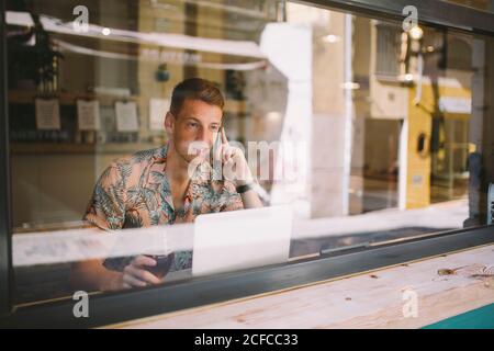 Focused young man talking on smartphone while using laptop at table throughout a windows in coffee shop Stock Photo
