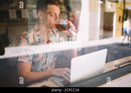 Focused young man talking on smartphone while using laptop at table throughout a windows drinking beer in coffee shop Stock Photo