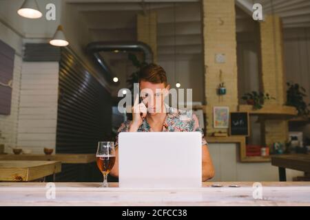 Concentrated man talking on smartphone while using laptop at table with drink in kitchen Stock Photo