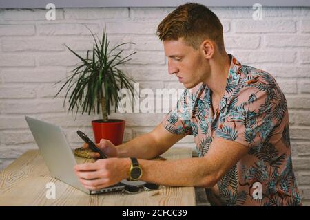Concentrated man talking on smartphone while using laptop at table with drink in coffee shop Stock Photo
