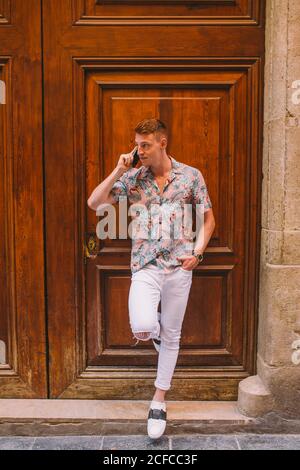 Handsome man talking on smartphone and leaning on wooden door looking away with hand in pocket Stock Photo