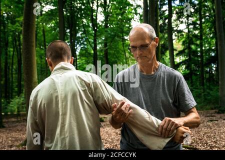 Bald mature instructor showing exercise to young man in uniform while practicing martial arts on blurred background of forest Stock Photo