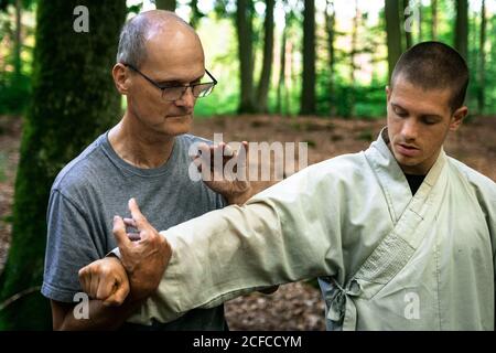 Bald mature instructor showing exercise to young man in uniform while practicing martial arts on blurred background of forest Stock Photo