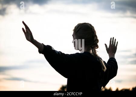 Silhouette of lady raising arms and meditating against sundown sky during workout in countryside Stock Photo