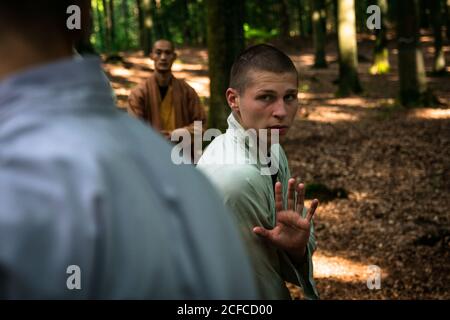 Bald mature instructor showing exercise to young man in uniform while practicing martial arts on blurred background of forest Stock Photo