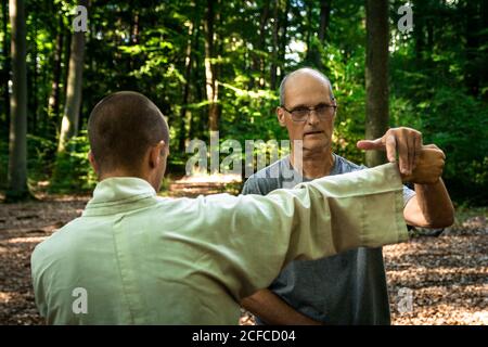 Bald mature instructor showing exercise to young man in uniform while practicing martial arts on blurred background of forest Stock Photo