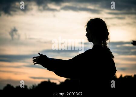 Silhouette of lady raising arms and meditating against sundown sky during workout in countryside Stock Photo