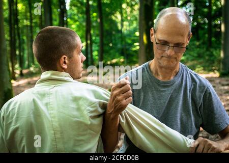 Bald mature instructor showing exercise to young man in uniform while practicing martial arts on blurred background of forest Stock Photo