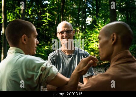 Bald mature instructor showing exercise to young man in uniform while practicing martial arts on blurred background of forest Stock Photo