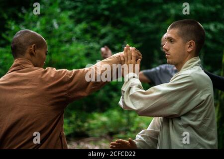 Bald mature instructor showing exercise to young man in uniform while practicing martial arts on blurred background of forest Stock Photo