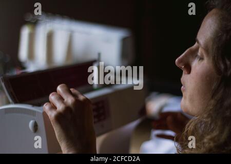 Serious young Woman using sewing machine making clothes working on dress sitting at table in house Stock Photo