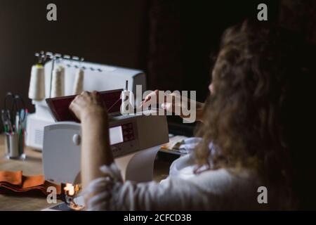 Back view of anonymous crop Woman using sewing machine making clothes sitting at table in house Stock Photo