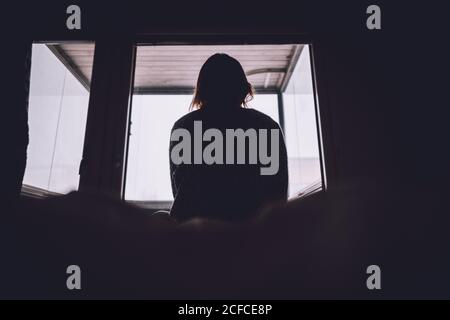 From bellow silhouette of anonymous lonely Woman sitting on bed against window in dark room at home Stock Photo