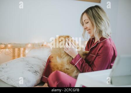 Young happy smiling attractive Woman in pajamas sitting in bed with little fluffy dog Stock Photo