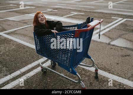 Smiling Woman sitting in shopping trolley in parking lot Stock Photo