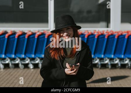 Attractive young Woman with red hair in black hat and jacket smiling with mobile phone and looking at camera in parking lot Stock Photo
