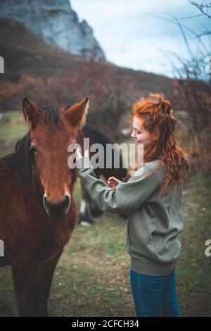 Side view of smiling Woman with long red hair in hoodie stroking brown horse with black mane in mountain pasture Stock Photo