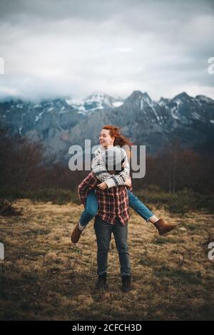 Lovely couple hugging and spending time together piggybacking and traveling in lawn with dry grass near by mountains in cloudy day Stock Photo