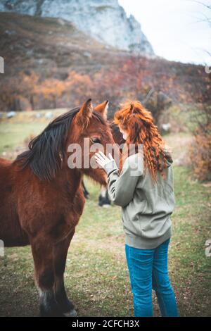 Side view of smiling Woman with long red hair in hoodie stroking brown horse with black mane in mountain pasture Stock Photo