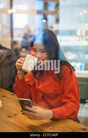 Young asian Woman using mobile phone while having cup of coffee at table in cafe Stock Photo