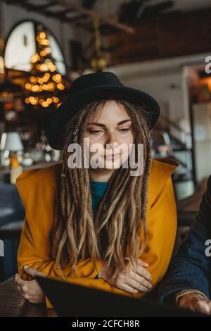 Smart man in glasses and stylish Woman in hat looking at monitor while man typing on laptop in cafe Stock Photo