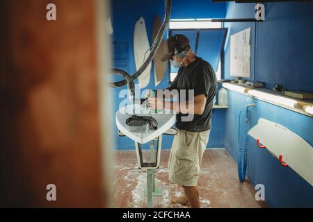 Side view of qualified worker in protective mask handling white surfboard in workshop with blue walls Stock Photo