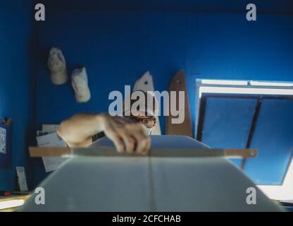 Concentrated qualified worker in protective mask aligning white surfboard in workshop with blue walls Stock Photo