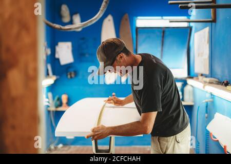 Side view of adult man measuring white board while working in small workshop with blue walls and making surf board Stock Photo