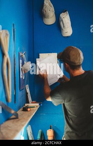 Back view of craftsman holding in hand blueprints of surf board while standing in workshop next to blue wall with tools Stock Photo