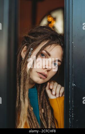 Stylish millennial female with dreadlocks wearing yellow coat over blue sweater looking away thoughtfully while sitting in room with piano in background Stock Photo