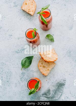 From above of glass bottles with fresh cold gazpacho soup on rough gray surface with toasts and basil Stock Photo