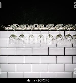 Row of clean polished wineglasses hanging against white tiled wall in vintage bar Stock Photo
