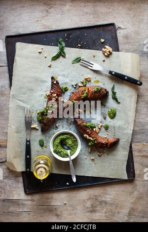 Top view of delicious sweet potato with fresh pesto sauce placed on tray and paper near forks and oil on wooden table Stock Photo