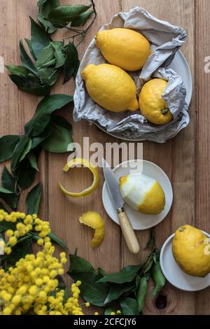 Top view of peeled and fresh lemons on plates on wooden table with green leaves and yellow flowers Stock Photo