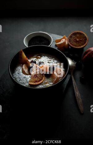 From above chia seeds pudding with slices of orange, mandarins and olives in bowl near spoon and grapefruit on dark table Stock Photo