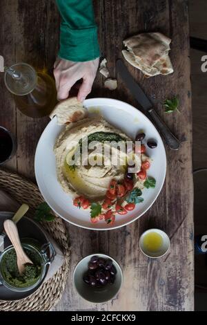 From above hand of anonymous person dipping piece of fresh flatbread into tasty hummus pesto decorated with cherry tomatoes and beans with parsley over lumber tabletop Stock Photo