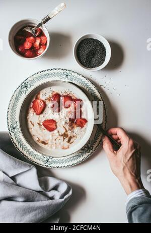 From above crop hand of man holding spoon for eating served oatmeal with strawberry and chia seeds Stock Photo