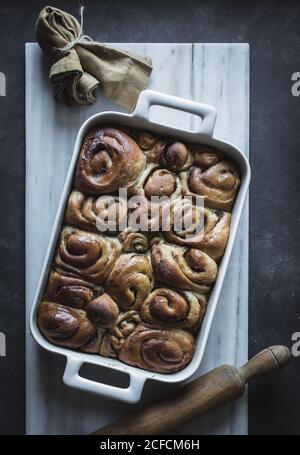 From above tray with fresh homemade cinnamon buns and wooden rolling pin on white marble board Stock Photo