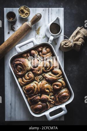 From above tray with fresh homemade cinnamon buns and wooden rolling pin on white marble board Stock Photo
