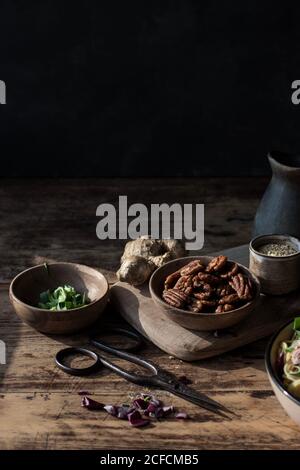 Bowl with pecan nuts near scissors, cutting board, pitcher and ginger in darkness Stock Photo