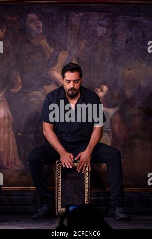 Bearded Hispanic guy playing percussion while sitting against mural during flamenco performance Stock Photo