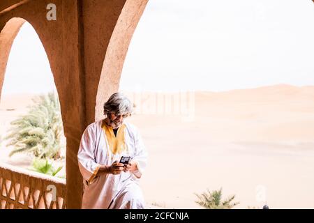 Side view of adult man in long outfit using phone standing on stone balcony with desert in the background, Morocco Stock Photo