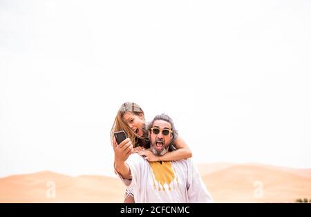 Middle-aged man with Woman piggyback taking selfie expressively on terrace against sandy desert, Morocco Stock Photo
