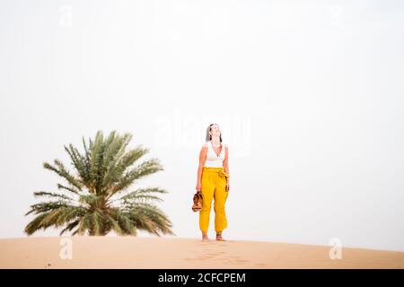 Cheerful stylish blonde Woman holding shoes while walking in desert of Morocco Stock Photo