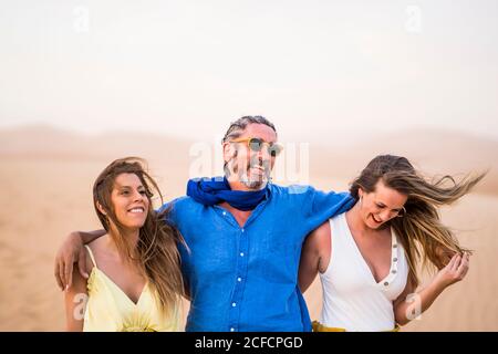 Senior bearded man laughing and hugging cheerful women while walking in sandy desert during trip in Morocco Stock Photo