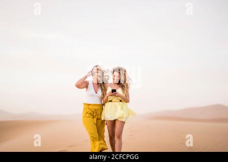 Cheerful stylish two blonde women friends using mobile phone while walking in desert of Morocco Stock Photo